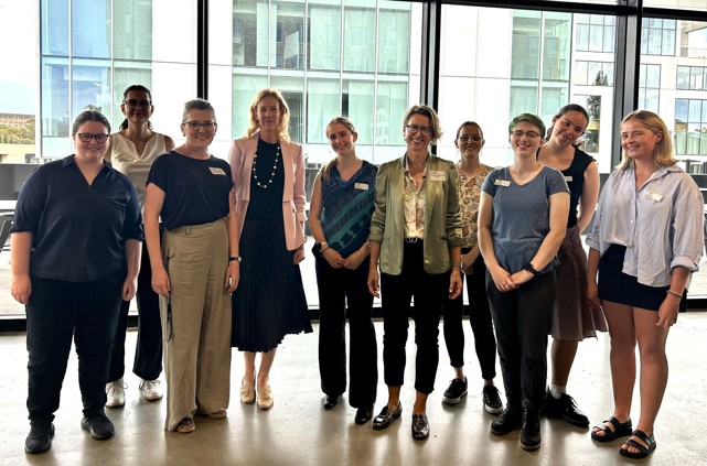 A group of ten women, including 8 young university students standing in a brightly lit room in front of a large window.
