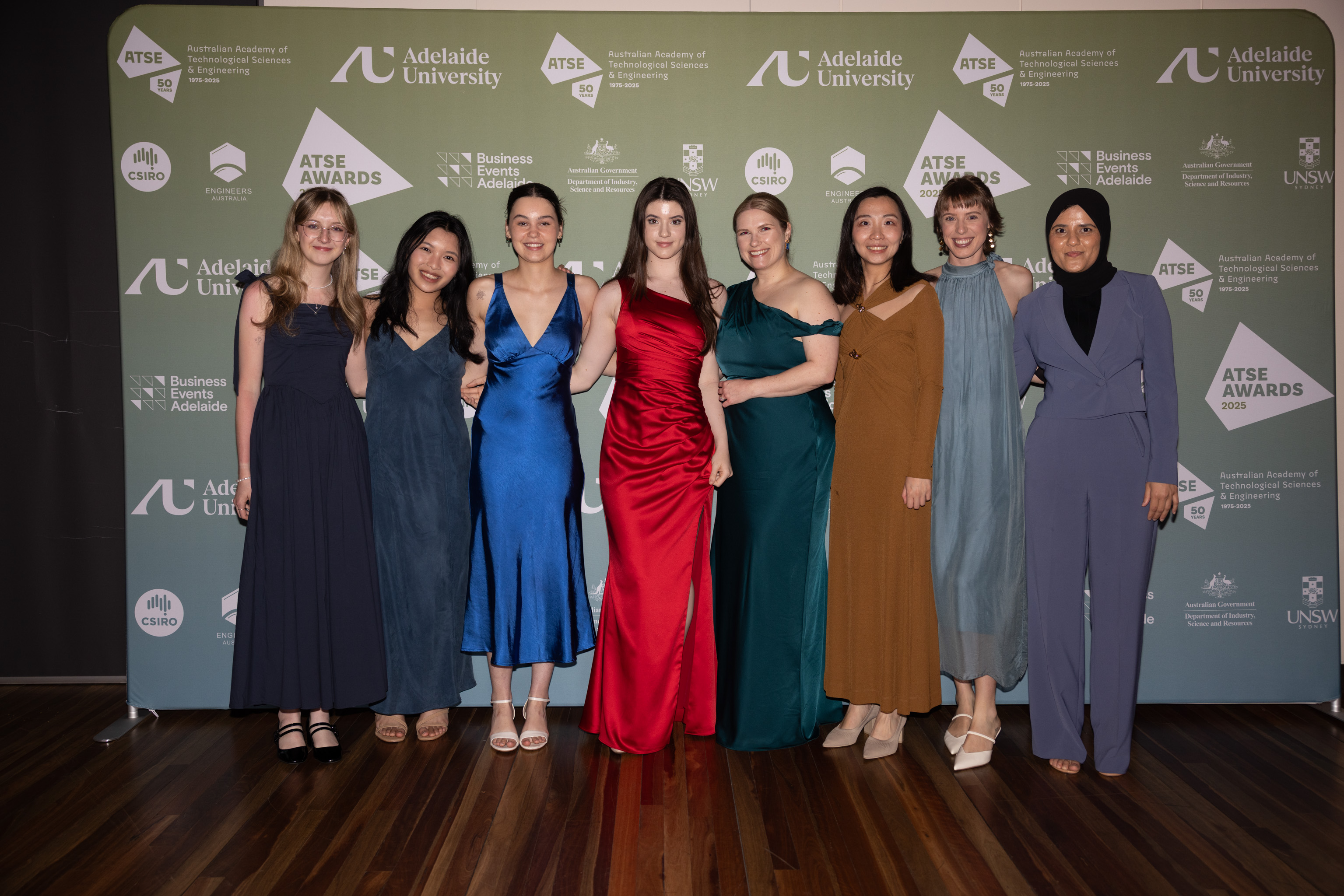 Eight young women in cocktail outfits standing together in front of a big media wall with logos.
