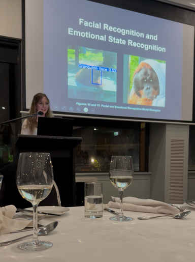 A young woman presenting at a lectern with a slide behind her on "Facial recognition and emotional state recognition" with pictures of orangutans.